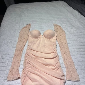 Champagnefärgad kort klänning med pärlor - Supersnygg kort klänning i champagnefärg med vadderad byst och långa meshärmar dekorerade med pärlor och små glaspärlor. Klänningen har rynkade detaljer längs sidorna och en figurnära passform. Perfekt för dig som vill sticka ut med eleganta detaljer och unik design. Köpt från ohpolly 