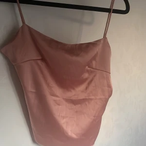 Rosa satinlinne med smala axelband - Säljer ett ljusrosa linne i satin med smala axelband och snygg drapering framtill. Toppen har en enkel knytning i ryggen och en lätt glansig finish som ger en lyxig känsla. Perfekt för dig som gillar en stilren och feminin look.