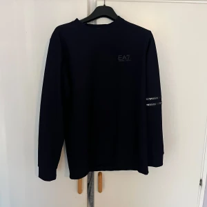 Blå långärmad tröja EA7 Armani - Blå långärmad tröja från EA7 Emporio Armani med diskret logga på bröstet och två svarta band med vit logotyp på ena ärmen. Tröjan har en clean och stilren look, perfekt för dig som gillar exklusiva streetwear-vibbar.