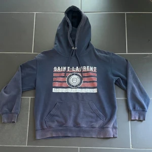 Marinblå Saint Laurent hoodie  - Snygg marinblå hoodie från Saint Laurent med vit och röd vintageinspirerad logga och tryck framtill. Tröjan har huva med snörning och en stor magficka. Såklart äkta. Storlek M men sitter som S 