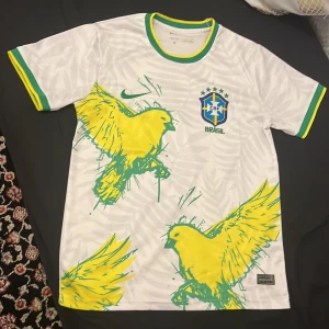 Vit Brasilien fotbollströja Nike S - Snygg vit Brasilien fotbollströja från Nike med gröna och gula detaljer. Använt 1 gång. Framsidan har stora gula fåglar och diskret bladmönster i bakgrunden. Broderat Brasilien-emblem och Nike-logga. Tillverkad i Dri-FIT material som andas.