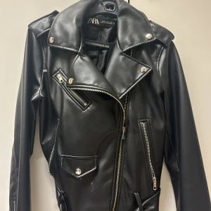 Svart skinnjacka från Zara - Snygg svart skinnjacka från Zara med klassisk bikerstil. Jackan har flera silvriga dragkedjor, stora slag och bälte i midjan. Perfekt för dig som vill ha en edgy look med coola detaljer och stilren passform.
