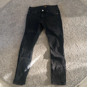 Svarta Lee Luke jeans W29 L30 - Snygga svarta jeans från Lee i modellen Luke. De har en smal passform med klassisk femficksdesign och raka ben. Jeansen är tillverkade i bomull med en stretchig känsla för extra komfort. Perfekta för dig som gillar en stilren och enkel look.