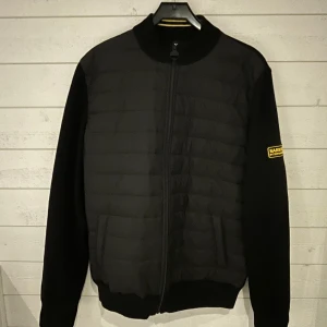 Svart cardigan från Barbour - Svart bomberjacka från Barbour International med quiltad front och ärmar i mjukare material. Jackan har ribbade muddar, hel dragkedja och en gul logotyp på ärmen. Perfekt för dig som gillar stilren och sportig look.