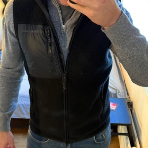 Svart fleeceväst från Peak Performance - Svart fleeceväst från Peak Performance med dragkedja framtill och en bröstficka i blankt material. Västen har en enkel, sportig look och är perfekt att slänga över en hoodie eller tröja. Snygg och funktionell för kyliga dagar.