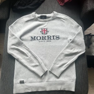 Grå stickad tröja från Morris - Snygg grå stickad tröja från Morris med broderad logga och brittisk flagga på bröstet. Tröjan har rund halsringning, ribbade muddar och nederkant samt lång ärm. Perfekt för dig som gillar klassisk och stilren design. Nypris ca 1100kr-1300kr
