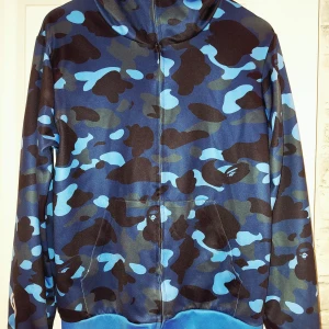 Blå camo zip up hoodie Bape - Säljer en ikonisk blå camo hoodie från Bape med dragkedja och huva. Tröjan har broderade patchar på huvan, ribbade muddar och klassiskt Bape-märke på ärmen. Stor M/liten L typ. 