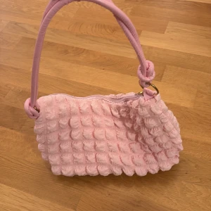 Rosa fluffig axelväska med struktur - Supergullig rosa axelväska med fluffig, strukturerad yta och mjukt material. Väskan har ett brett handtag med knutdetaljer och stängs med dragkedja. Perfekt för dig som vill sticka ut med en söt och trendig accessoar.