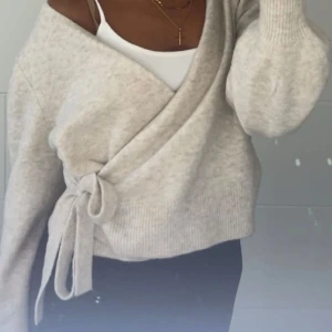 Beige omlottkofta med knyt - Superfin beige omlottkofta med knyt i sidan och v-ringad hals. Koftan är stickad och har långa ärmar med ribbade muddar. Perfekt att slänga på sig över ett linne för en chill och snygg look.