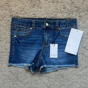 Blå jeansshorts med fransad kant från RYVLS - Snygga blå jeansshorts från RYVLS med fransad kant och klassiska fickor både fram och bak. Shortsen har guldiga knappar, normal passform och midwaist. Perfekta för sommarens alla äventyr, med slitna detaljer för en cool vibe.