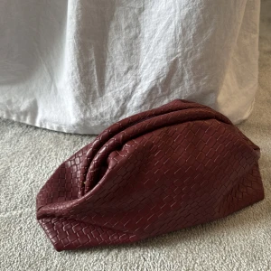 Pouch väska vinröd  - Säljer en vinröd pouch väska i flätat skinnimitation med cool kedjedetalj i guld. Väskan har en mjuk, slouchy form och lång axelrem. Perfekt för dig som vill ha en trendig och rymlig väska med unik textur.