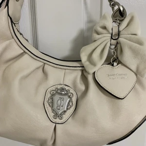 Vit axelväska Juicy Couture med rosett - Superfin vit axelväska från Juicy Couture i skinn med silverfärgad JC-logga framtill. Väskan har en kedjedetalj på axelremmen, en stor mjuk rosett och ett hjärtformat hänge. Svarta kontrastsömmar och snyggt rynkad form ger den en unik look.