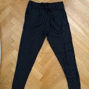 Svarta randiga byxor med snörning - Svarta stretchiga byxor med vita vertikala ränder och elastisk midja med snörning. Byxorna har fickor fram, samt en avslappnad passform som smalnar av nedtill. Perfekta för en skön och stilren look