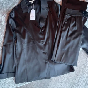 Svart kostym i satin från Sanne Alexandra - Supersnygg svart skjorta och byxor glansigt satinmaterial från Sanne Alexandra. Klassisk krage, långärmad och med knappar framtill. Ny med prislapp 