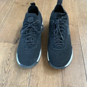 Svarta sneakers från  Hugo BOSS - Svarta sneakers från Hugo BOSS med stickad ovandel och vita detaljer på sulan. Skorna har snörning med svarta skosnören med vita prickar och en elastisk rem med BOSS-logga på sidan. Snygg och modern design med rund tå och platt sula. Storlek 45