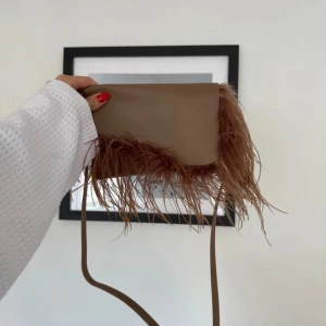 Brun axelväska med fjädrar från ATP - Säljer en brun axelväska från ATP i mjukt skinn med unika fjäderdetaljer längs kanterna. Väskan har ett smidigt axelband, magnetstängning och ett fack på baksidan. Perfekt för dig som vill sticka ut med en cool accessoar.