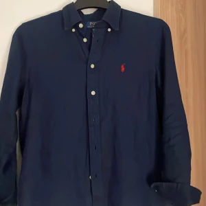 Mörkblå  linne skjorta från Polo Ralph Lauren - Klassisk mörkblå skjorta från Polo Ralph Lauren med vit knappstängning och röd broderad logga på bröstet. Endast använd några gånger och är i mycket bra skick. Nästan nyskick. Storlek:  M (10-12).  Nypris: 1100kr. Säljer då den var för liten, lägg gärna prisförslag.