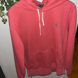 Rosa hoodie från Polo Ralph Lauren - Passar 170-185cm Snygg rosa hoodie från Polo Ralph Lauren med klassisk blå logga på bröstet. Tröjan har huva med vit snörning och en stor magficka framtill. Mjuk bomullskvalitet och långärmad modell, perfekt för chill dagar.