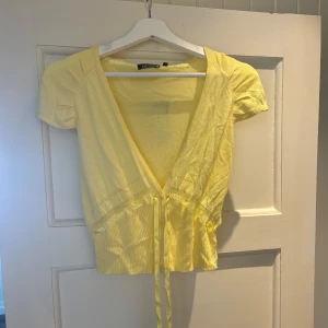 Gul stickad blus från Dulcie - Söt gul stickad blus från Dulcie med korta puffärmar, djup v-ringning och knappar framtill. Blusen har dekorativ snörning i midjan och fina stickade detaljer. Perfekt för en somrig look med sin ljusa färg och lätta material.