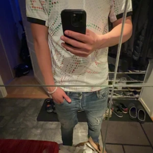 Portugal fotbollströja Puma vit - Säljer en Portugal fotbollströja från Puma i vitt med svarta detaljer och färgglada prickmönster i grönt och rött. Tröjan har korta ärmar, svart krage och Portugal-emblem på bröstet. Perfekt för dig som vill sticka ut på match eller träning.