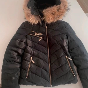 Svart pufferjacka med pälsluva - Svart pufferjacka med snygga guldfärgade dragkedjor och flera fickor. Jackan har en stor huva med fluffig beige päls och quiltad design. Långa ärmar med ribbad mudd på insidan för extra värme. Perfekt för kalla dagar och riktigt stilren look.