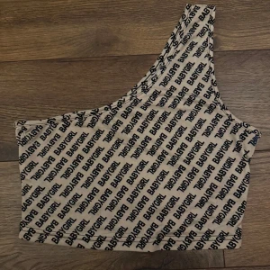 Beige one shoulder topp med BABYGIRL-tryck - Säljer en beige one shoulder topp med svart textmönster där det står BABYGIRL över hela. Toppens asymmetriska design gör den unik och den är tillverkad i ett mjukt, stretchigt material som sitter tight. Perfekt för dig som vill sticka ut med en trendig look.