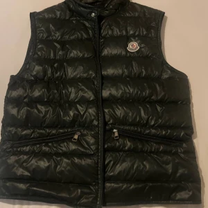 Svart dunväst från Moncler - 100% äka top skick Svart dunväst från Moncler som passar perfekt för lite kallet klimat. Västen har hög krage, två snedställda fickor med dragkedja och Moncler-logga på bröstet. Perfekt för lager-på-lager och stilren look. 