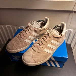 Adidas Gazelle rosa sneakers mocka - Säljer ett par klassiska Adidas Gazelle sneakers i rosa mocka med vita detaljer och de ikoniska tre ränderna på sidan. Skorna har snörning, platt sula och rund tå. Perfekta för dig som gillar stilrena och tidlösa sneakers med retrovibbar.