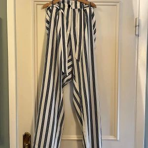 Randiga byxor med knyt från Lindex - Snygga vita byxor med svarta vertikala ränder från Lindex. Byxorna har hög midja, vida ben och ett matchande knytband i midjan. Perfekta för en chill och trendig look. Resår i midjan för extra komfort.