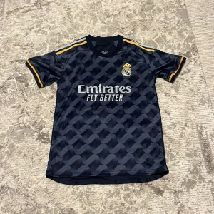 Real Madrid Vini Jr. fotbollströja #7 - Snygg Real Madrid fotbollströja med Vini Jr. och nummer 7 på ryggen. Tröjan är mörkblå med ett vågigt mönster, gula detaljer på axlar och ärmslut samt klubbmärke på bröstet. Tillverkad i lätt polyester, perfekt för du som älskar Real Madrid.