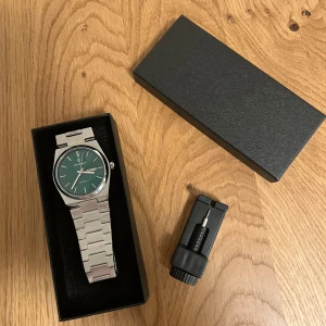 Grön och silverfärgad klocka från Poedagar  - Stilren klocka  med grön urtavla och dag/datumvisning. Armband och boett i silverfärgat metall, klassisk design med raka länkar. Levereras i svart ask och inkluderar verktyg för att justera armbandet. Perfekt klocka för en modern look.