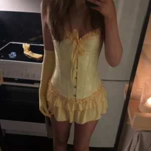 Gul korsett med kjol med volanger - Supersöt gul korsett med kjol  med korsettliv, snörning i ryggen och volangkant. Axelbandslös modell med söt rosett framtill och matchande långa handskar. Klänningen har en figurnära passform. Man får med handskar och tiara använt en gång💕kjolen har inbyggda shorts 