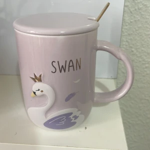Mugg  - Bilderna visar en ljuslila mugg med lock och sked, dekorerad med en svan och texten 'SWAN'. Muggen är i keramik och ser ut att vara i mycket gott skick utan synliga defekter eller slitage. Perfekt för te eller kaffe. Ej elektronik.