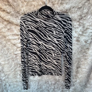 Zebramönstrad polotröja i svart och beige - Snygg polotröja med zebramönster i svart och beige. Tröjan har lång ärm och en tight passform med hög krage. Skönt material. Lappar saknas. B 37 cm, L 54 cm