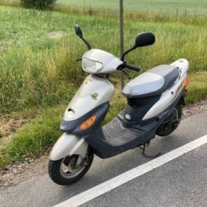 Baotian classic delar  - Bilden visar en grå moped/scooter parkerad vid vägkanten. Mopeden ser ut att vara i bruksskick med synligt slitage på fotplattan och vissa repor på kåporna. Inga uppenbara defekter syns, men produkten är inte elektronik och matchar inte någon av de angivna taxonomierna.