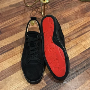 Christian Louboutin svarta sneakers - Snygga svarta sneakers från Christian Louboutin i mocka med klassisk röd sula. Skorna har rund tå, svarta skosnören och diskret branding på insidan. Perfekta för dig som vill ha en clean och lyxig look med en ikonisk touch. Kommer med kvitto!