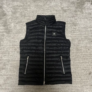 Moose Knuckles väst - Svart quiltad dunväst från Moose Knuckles med hög krage och två dragkedjeförsedda fickor. Västen har en stilren look med logotyp på bröstet och silverfärgad dragkedja framtill. Perfekt för lager-på-lager under kyliga dagar.