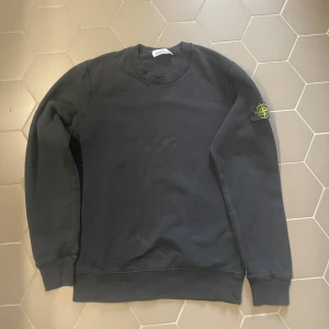 Svart sweatshirt från Stone Island - Svart sweatshirt från Stone Island med klassisk rund hals och ribbade muddar. Tröjan har den ikoniska loggan på vänster ärm. Tillverkad i mjuk bomull och har en normal passform. Perfekt för dig som gillar stilrena och snygga streetwear-plagg. KAN FÖRHANDLA OM PRISET!