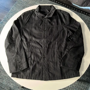 Svart jacka från a.v.v HOMME, stl 46 - Svart jacka från a.v.v HOMME i storlek 46. Klassisk design med knappar framtill, krage och fickor med lock. Långärmad modell i bomullsmaterial, perfekt för dig som gillar stilren och enkel look.