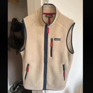 Patagonia vest  - Säljer en beige fleeceväst från Patagonia med blå detaljer och röda dragkedjor. Västen har hög krage, tre fickor med dragkedja. Perfekt för lager-på-lager och outdoor-vibe.
