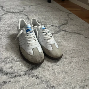 Adidas Samba vita sneakers - Klassiska Adidas Samba sneakers i vitt med grå mockadetaljer och bruna gummisulor. Skorna har de ikoniska tre ränderna på sidan och blå Adidas-logga på plösen. Perfekta för dig som gillar retrostil och streetwear.