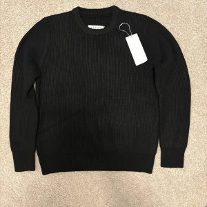 Svart stickad tröja från Maison Margiela - Säljer en svart, ribbstickad tröja från Maison Margiela. Tröjan har rund halsringning, långa ärmar och klassiska fyra vita stygn i nacken. Perfekt för dig som gillar minimalistisk och stilren design.