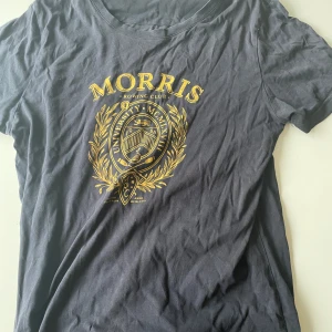 Svart t-shirt från Morris R. Club - Svart t-shirt från Morris R. Club med stort guldfärgat tryck framtill i college-stil. Klassisk rund hals och korta ärmar. Skön passform och tillverkad i mjuk bomull. Perfekt för dig som gillar preppy och sportig vibe.