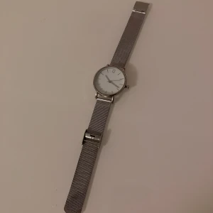 Silverfärgad klocka i rostfritt stål - Stilren klocka med rund urtavla och mesh-armband i rostfritt stål. Urtavlan är vit med minimalistiska markeringar och silverfärgade visare. Spännet är märkt med 'STAINLESS STEEL' och hela klockan har en modern, clean look. Behöver ett nytt batteri.