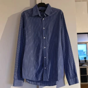 Blå randig skjorta Tiger of Sweden - Snygg blå skjorta från Tiger of Sweden med tunna vita ränder. Klassisk krage, knäppning framtill och långa ärmar med manschett. Tillverkad i mjuk bomull för en clean och stilren look. Perfekt för dig som gillar en tidlös vibe.