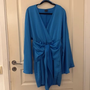 Blå omlottklänning med rosett - Supersnygg blå omlottklänning från Gina tricot  med lång ärm och stor rosett i midjan. Klänningen har v-ringning och är tillverkad i ett glansigt, mjukt material som ger en lyxig känsla. Perfekt för dig som vill sticka ut med färg och stil.