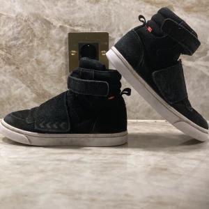 Svarta Hummel sneakers strl 33 - Svarta höga sneakers från Hummel i mocka med breda kardborreband och vit sula. Klassisk Hummel-logga på sidan och på remmen. Snygg och sportig modell med rund tå och vadderad plös för extra komfort.