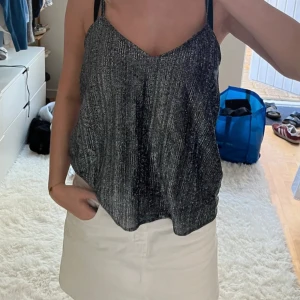 Glittrig svart linnetopp med smala axelband - Säljer en svart glittrig linnetopp med smala axelband och v-ringning. Toppen har ett lätt glansigt material och ett subtilt randigt mönster som ger en snygg effekt. Perfekt för dig som vill sticka ut lite extra med en enkel men cool topp.