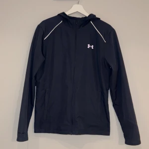 Svart vindjacka från Under Armour - Svart vindjacka från Under Armour och Jackan har riflex detaljer vid axlarna och stor logga på ryggen perfekt för aktiva dagar. Snygg och sportig look jackan är mycket ovanlig/selsynt obs möts endast 