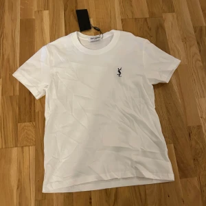 Vit Saint Laurent t-shirt med logga - Clean vit t-shirt från Saint Laurent med klassisk svart YSL-logga broderad på bröstet. T-shirten har rund halsringning och korta ärmar. Tillverkad i mjuk bomull för en skön känsla och stilren look.